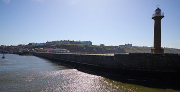 The image shows a landscape photograph of Whitby, Yorkshire, taken from the pier during the afternoon in the spring. In the foreground, the harbour is visible with the iconic Whitby lighthouse standing prominently on the right side of the pier, which stretches out into the water. Along the shoreline, residential and commercial buildings can be seen clustered together, with the higher elevation in the background offering views of larger establishments overlooking the town. The sunlight reflects off the water, adding brightness to the overall scene, and the weather is clear, highlighting the seasonal vibrancy of spring in this picturesque Yorkshire coastal location.